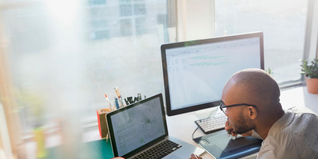 Image shows a man wearing glasses and working at two computer screens.