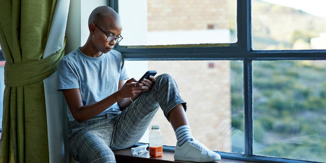 A woman sits on a windowsill overlooking greenery. She focuses on a cellphone, which she has in hand.