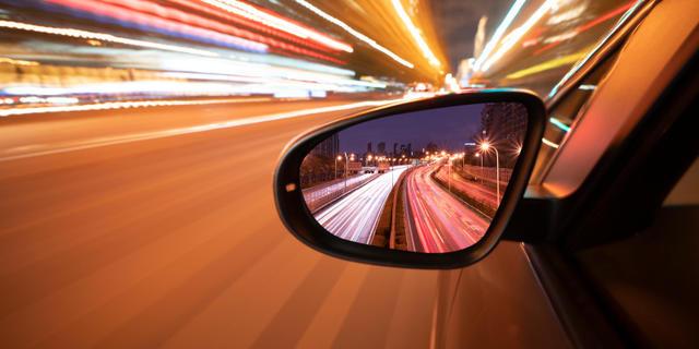 Side view mirror of car accelerating with blurred lights in the background.